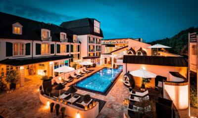 Luxurious hotel courtyard at dusk, featuring a vibrant blue-lit pool surrounded by white umbrellas and cozy lounge chairs, exuding a tranquil ambiance.