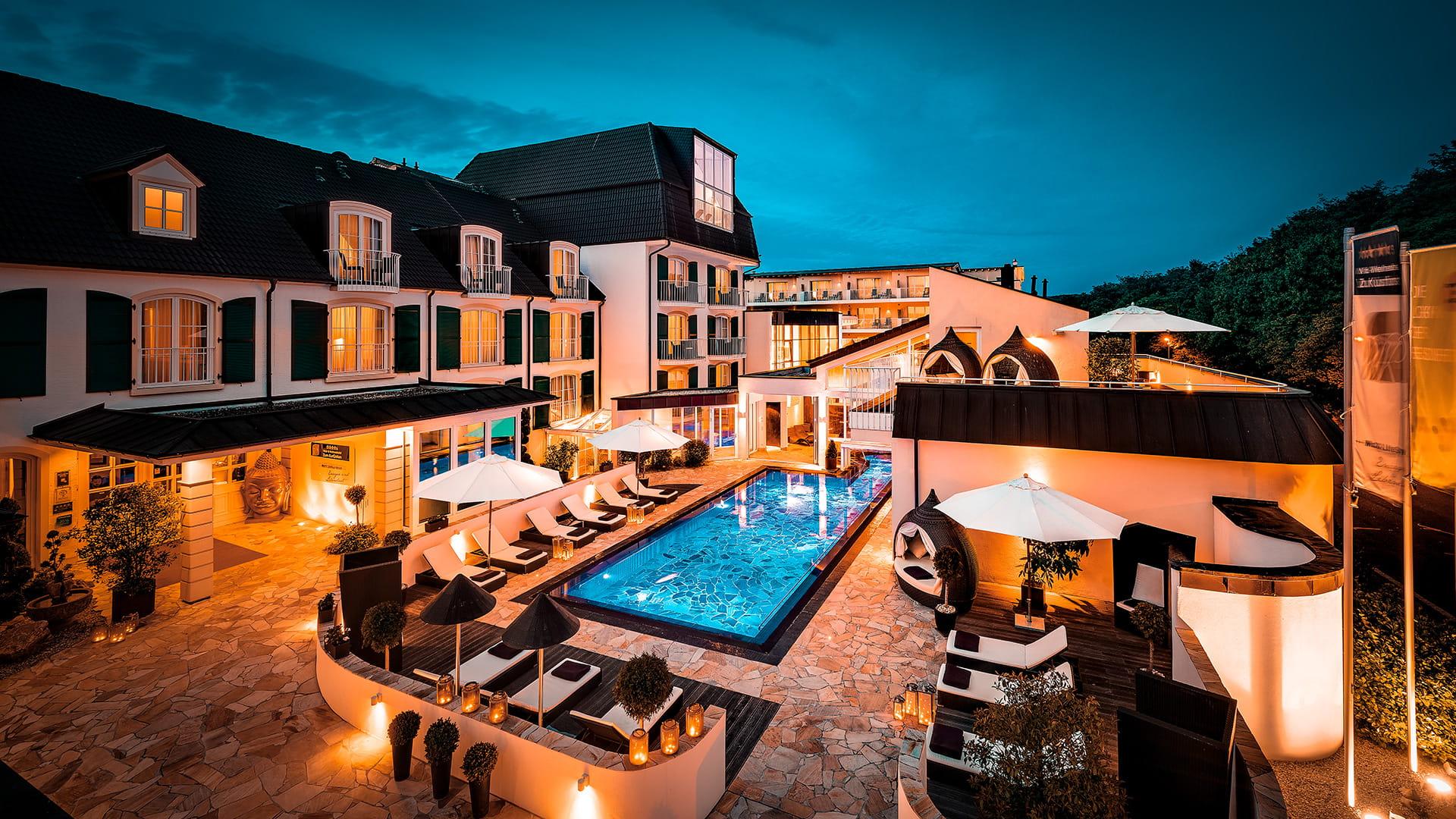 Luxurious hotel courtyard at dusk, featuring a vibrant blue-lit pool surrounded by white umbrellas and cozy lounge chairs, exuding a tranquil ambiance.