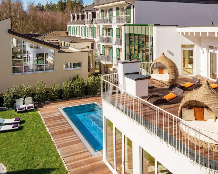 A view of the modern lifestyle resort Zum Kurfürsten, featuring several buildings, a terrace with wicker chairs, and a small pool on a wooden deck next to a sunbathing lawn
