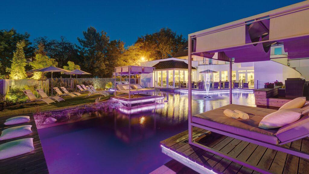 Nighttime view of an illuminated natural swimming pond with lounge chairs and covered lounge beds at the Lifestyle Resort Zum Kurfürsten in Bernkastel-Kues