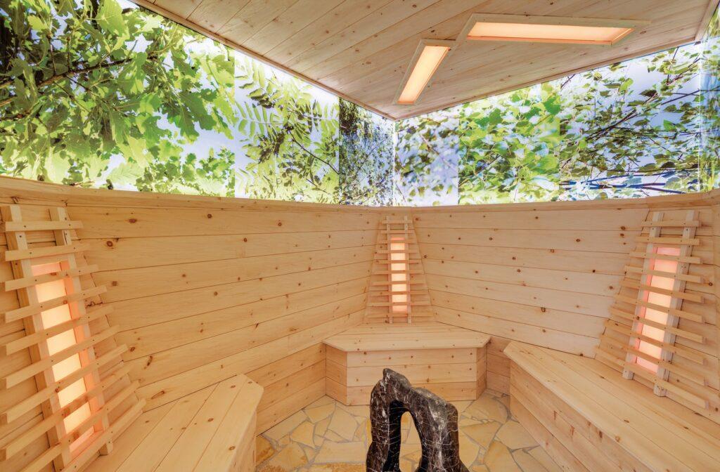 Sauna room with light-colored wooden benches and walls, indirect lighting, and large windows offering a view of the green treetops