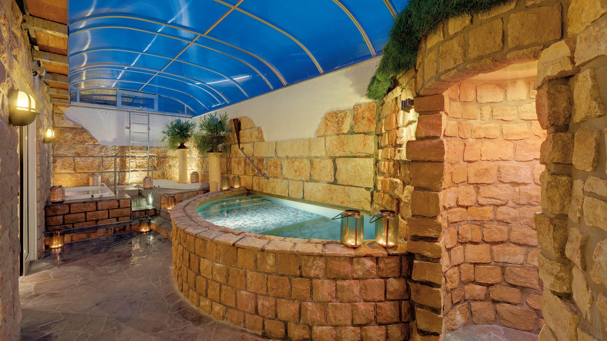 The interior of the wellness area at the Lifestyle Resort Zum Kurfürsten, featuring a small pool with natural stone masonry, a rock grotto, and a transparent roof