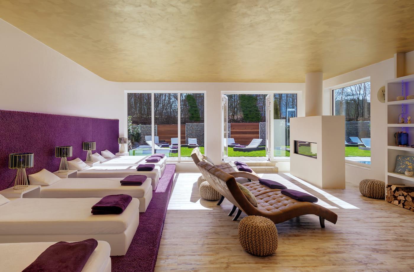Luxurious spa lounge with white loungers and purple accents, featuring a fireplace, large windows with garden views, and a warm, serene atmosphere.