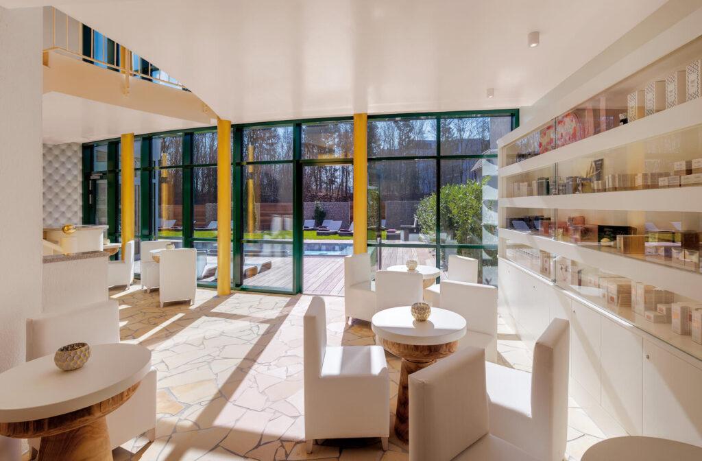 A bright wellness area with round wooden tables, white armchairs, large glass walls, and shelves stocked with products