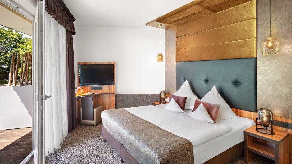 A modern hotel room at the Zum Kurfürsten Lifestyle Resort on the Moselle, featuring a large double bed, an upholstered headboard, two nightstands with lamps, a flat-screen TV, and access to a balcony with a view of the surrounding greenery