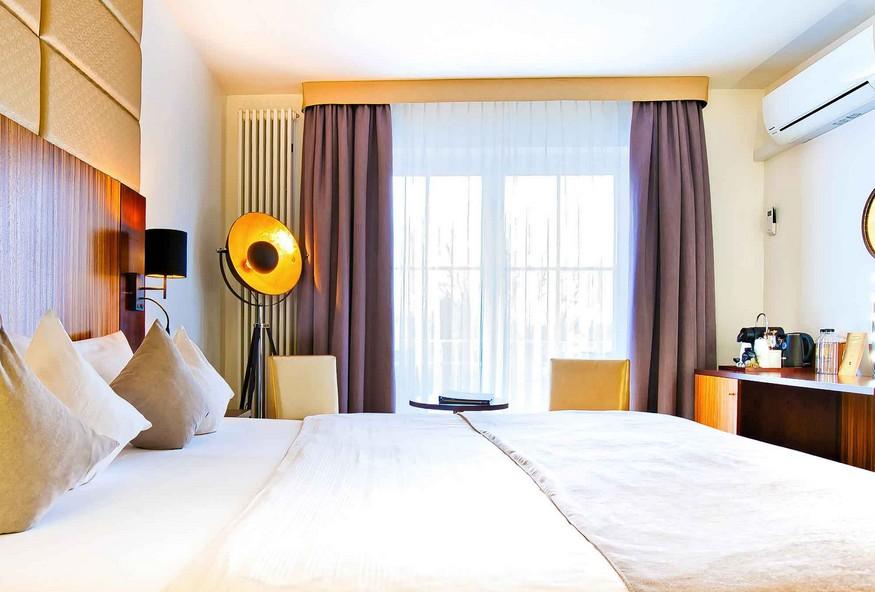 A modern hotel room at the Zum Kurfürsten Lifestyle Resort on the Moselle, featuring a double bed, a floor lamp with a large gold-colored shade, two chairs in front of floor-to-ceiling windows with curtains, and a desk with a coffee maker