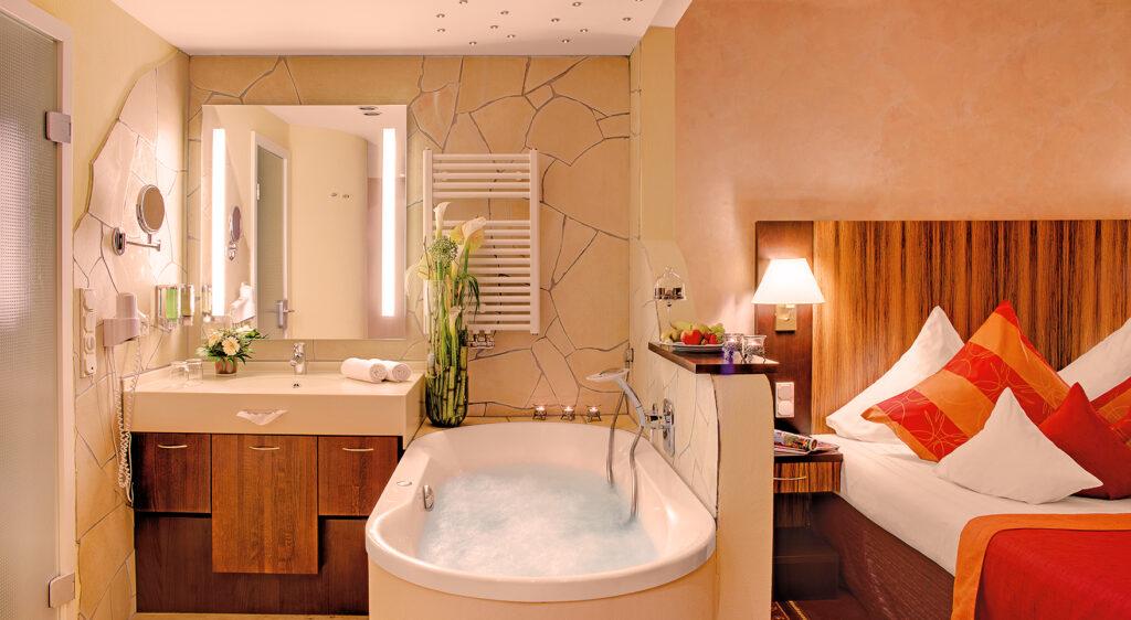 A hotel room at the Lifestyle Resort Zum Kurfürsten, featuring a bathtub, sink, towels, and decorative pillows on the bed