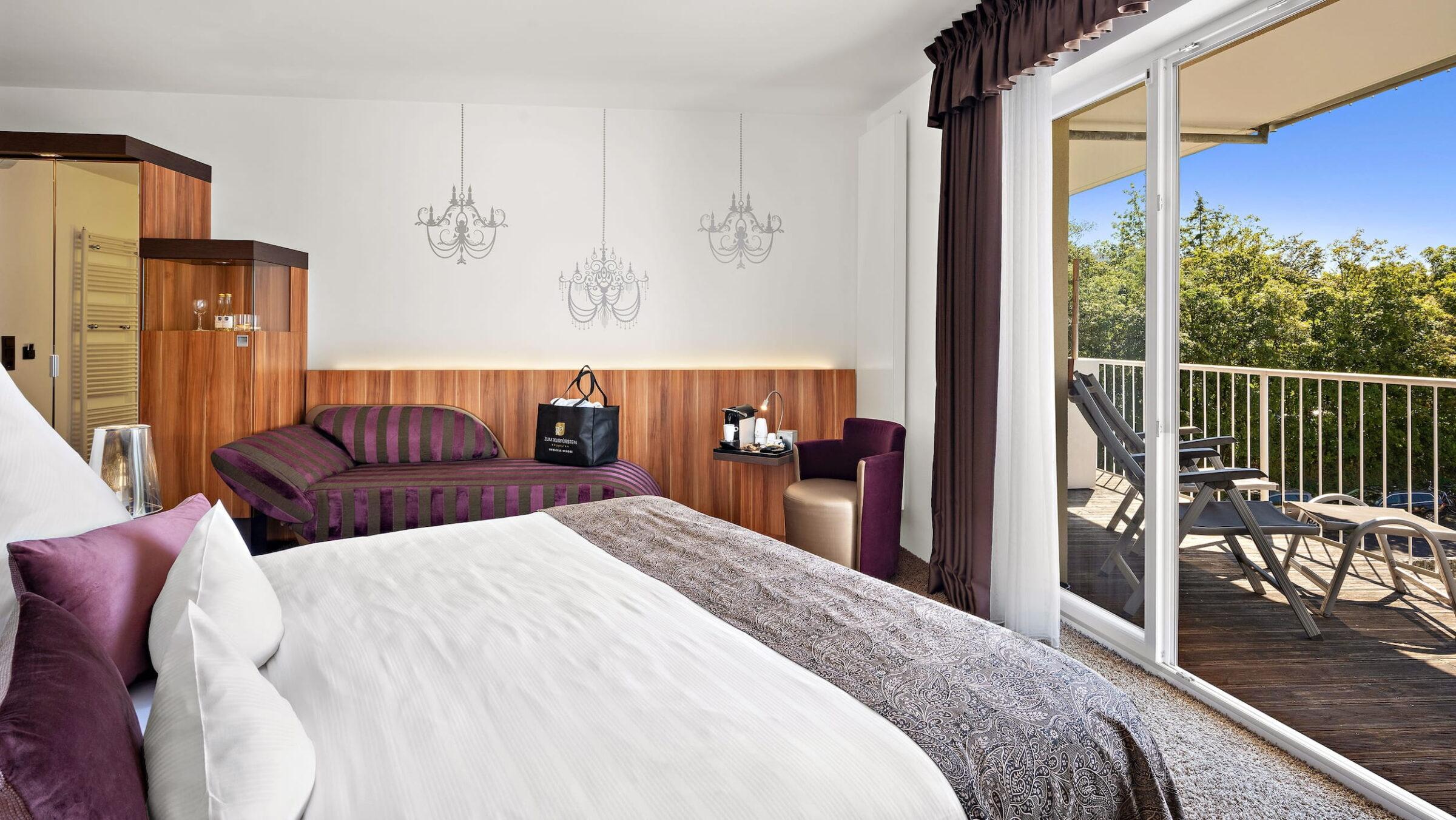 A hotel room at the Lifestyle Resort Zum Kurfürsten in Bernkastel-Kues featuring a double bed, a sitting area with a sofa and armchair, a wooden wall adorned with three chandeliers, and a balcony with lounge chairs and a view of green trees