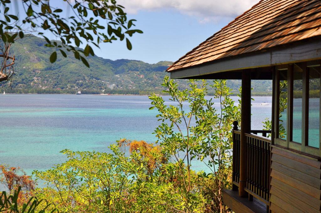 Blick von einer Holzvilla mit Schindeldach auf das türkisfarbene Meer und bewachsene Hügel im Hintergrund auf Cerf Island, Seychellen