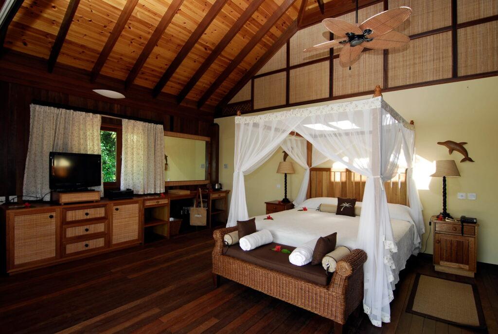 Innenraum einer nachhaltigen Villa im Cerf Island Resort auf den Seychellen mit Holzboden, Himmelbett mit Moskitonetz, Deckenventilator aus geflochtenem Material, Holzmöbeln und Vorhängen an den Fenstern