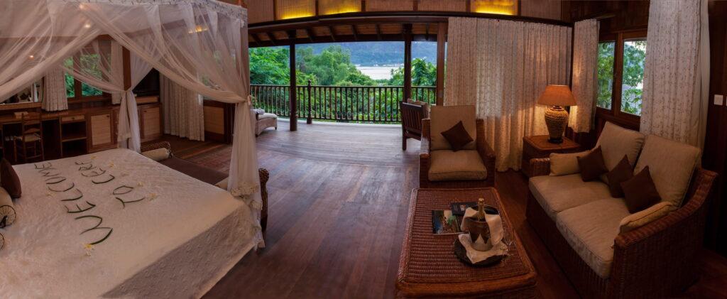 Innenraum einer nachhaltigen Hillside-Villa im Cerf Island Resort auf den Seychellen mit offenem Grundriss, Holzboden, Bett mit Moskitonetz, Sitzbereich mit Sofas und Blick auf tropische Vegetation und Meer