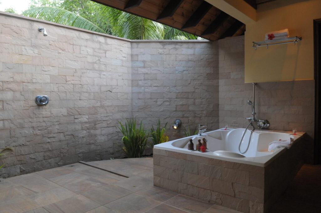 Freistehende Badewanne mit Armaturen und Duschkopf in einem halb offenen Badezimmer mit Steinmauer und Pflanzen im nachhaltigen Cerf Island Resort auf den Seychellen.