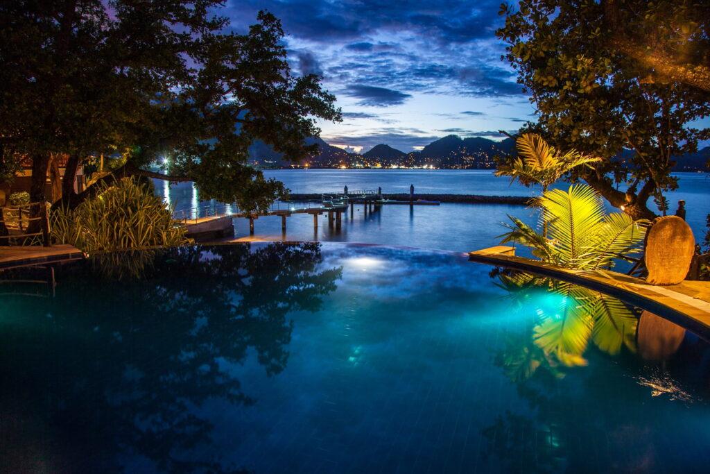 Rundlicher beleuchteter Pool in der Nacht mit Blick auf eine Bucht und Berge im Hintergrund umgeben von Bäumen und Pflanzen im nachhaltigen Cerf Island Resort auf den Seychellen