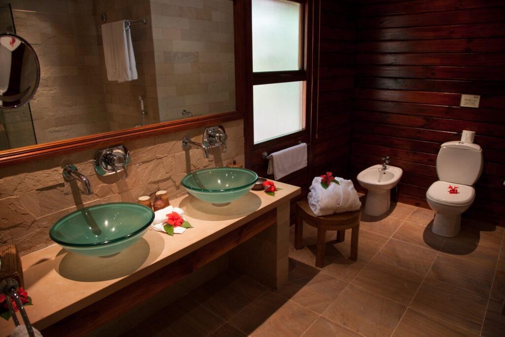 Badezimmer in ther Tortoise Suite des nachhaltigen Seychellen-Resorts auf Cerf Island mit zwei grünen Glaswaschbecken auf hellem Waschtisch, Holzwand, Toilette, Bidet, Handtüchern und roten Blüten als Dekoration.