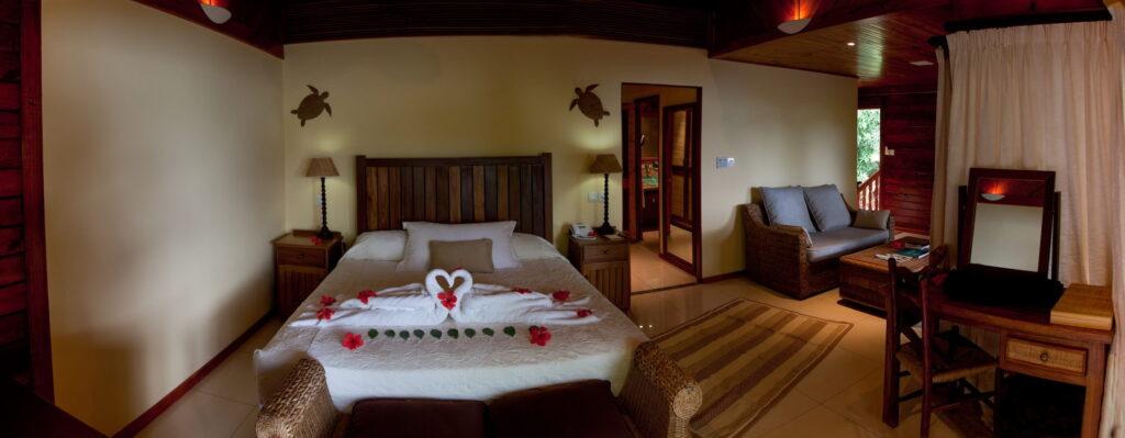 Schlafzimmer in der Tortoise Suite im nachhaltigen Cerf Island Resort mit Doppelbett, auf dem zwei Handtücher zu einem Schwan geformt sind, zwei Wandlampen in Schildkrötenform, Sofa, Schreibtisch mit Spiegel und Teppichläufer auf dem Boden