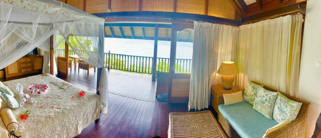 Offenes Schlafzimmer mit Himmelbett, Sofa, Teppich und Blick auf eine Holzterrasse mit Meerblick im nachhaltigen Cerf Island Resort auf den Seychellen