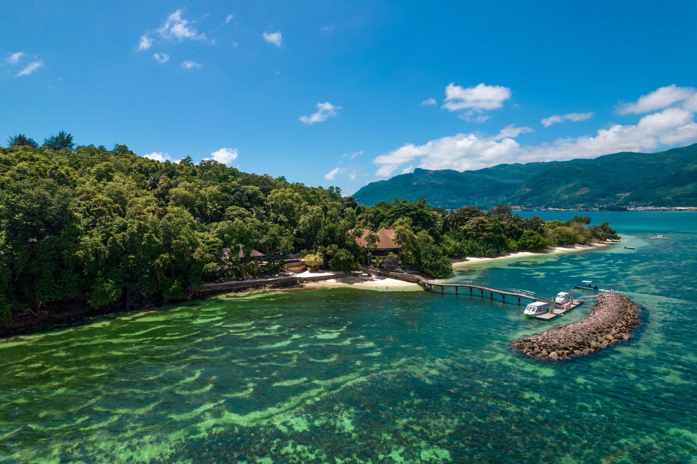Holzsteg führt von einem bewaldeten Strandabschnitt des Cerf Island Resorts auf den Seychellen zu einem kleinen Bootsanleger im türkisfarbenen Meer