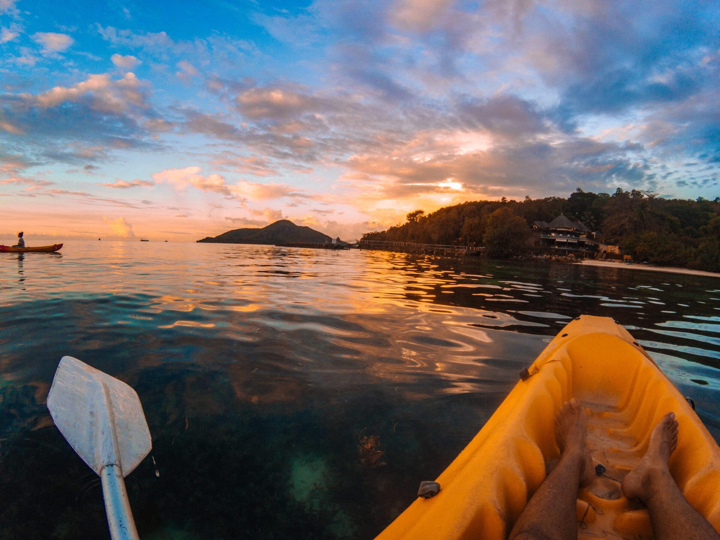 Blick von einem gelben Kajak auf ruhiges Meer mit Person im Kajak und bewaldeter Küste des Cerf Island Resorts bei Sonnenuntergang