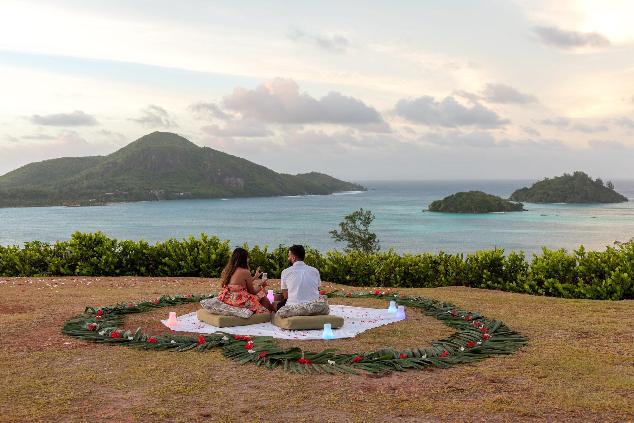 Paar sitzt auf einer Picknickdecke mit grüner Blätter- und roten Blütenumrandung auf einer Wiese mit Blick auf das Meer und bewachsene Inseln der Seychellen im Hintergrund