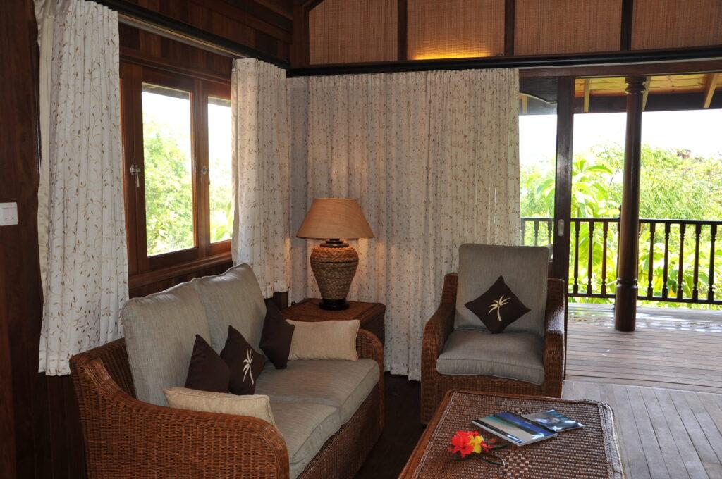 Sitzbereich mit Korbsesseln, Sofas, Kissen mit Palmenmotiv, Tisch mit Büchern und Blume vor großen Fenstern und Zugang zu einer Holzterrasse mit Blick auf tropische Vegetation im nachhaltigen Cerf Island Resort auf den Seychellen
