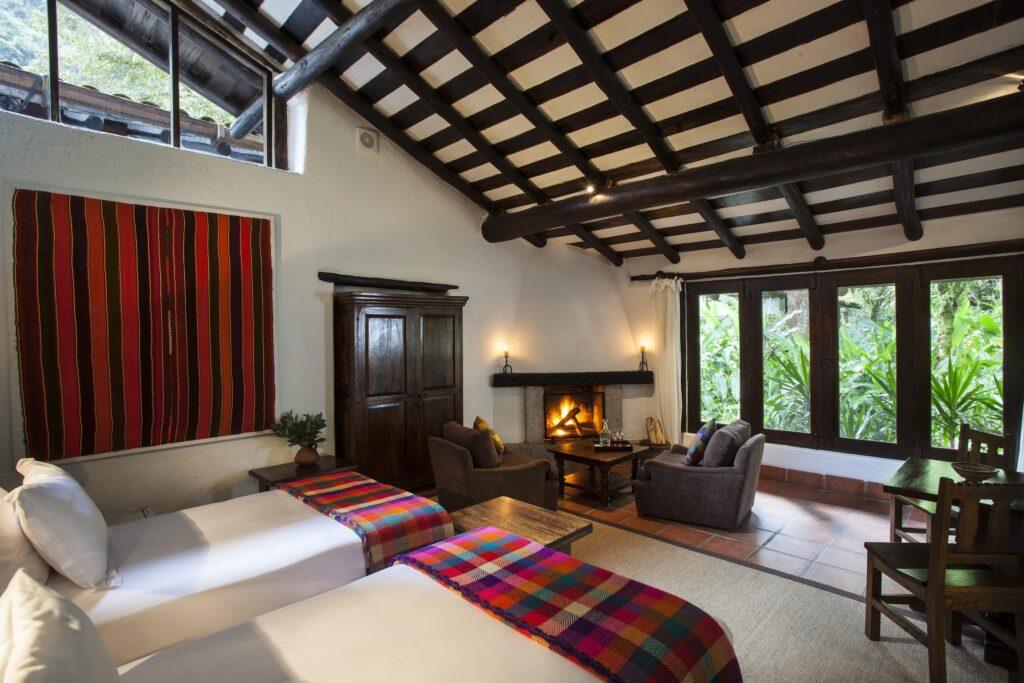 Gemütliches Zimmer im nachhaltigen Inkaterra Machu Picchu Pueblo Hotel mit zwei Betten, die mit bunten gestreiften Decken bedeckt sind, einem Holzschrank und einem beleuchteten Kamin. Große Fenster geben den Blick auf üppiges Grün nach draußen frei.
