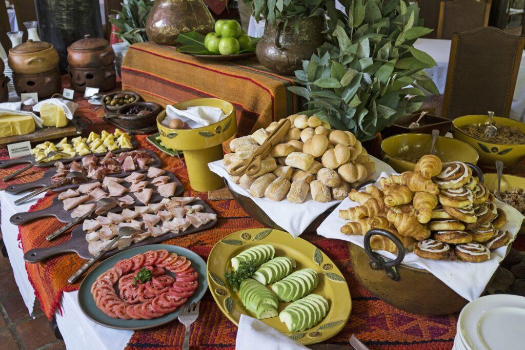 Buffettisch im Inkaterra Machu Picchu Pueblo Hotel mit einer Auswahl an Speisen: Aufschnitt, Brot, Gebäck, Tomaten, Avocados, Äpfel und dekorative Pflanzen. Warme, einladende Atmosphäre.