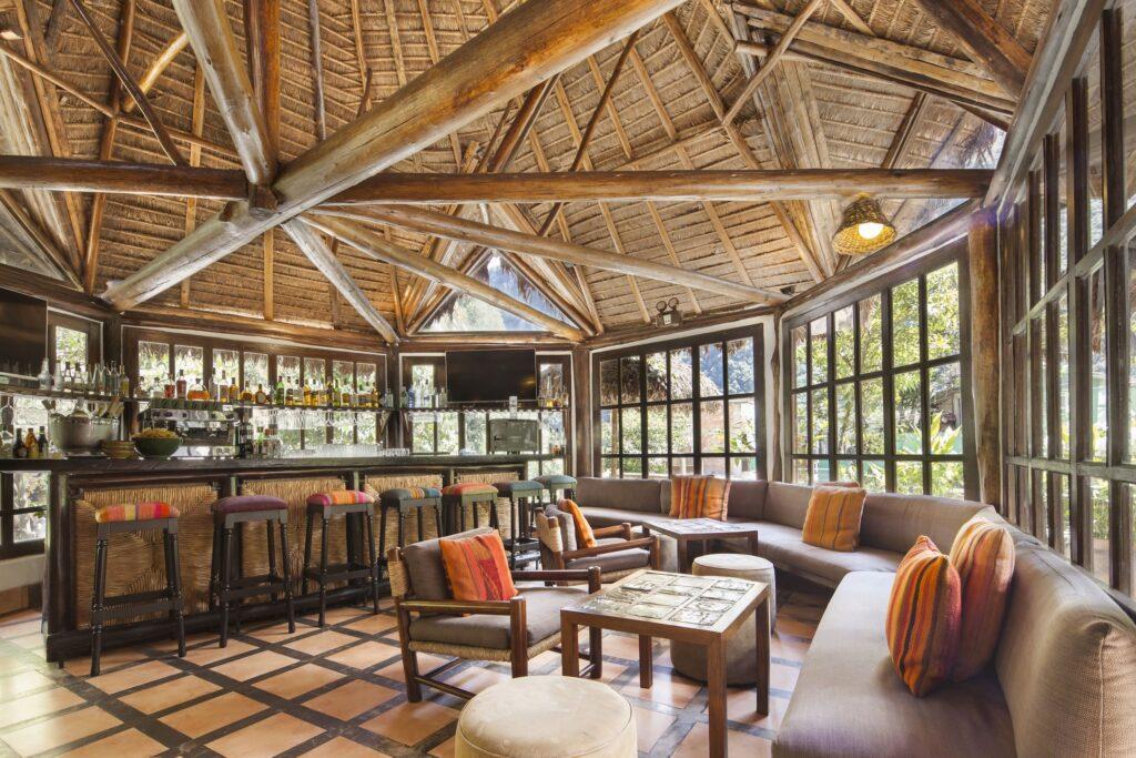 Rustikale Lounge im Inkaterra Café in Machu Picchu Pueblo mit Holzbalken, gemütlichen Sitzgelegenheiten, bunten Kissen und großen Fenstern. Eine Bar mit Hockern sorgt für eine einladende und entspannte Atmosphäre.