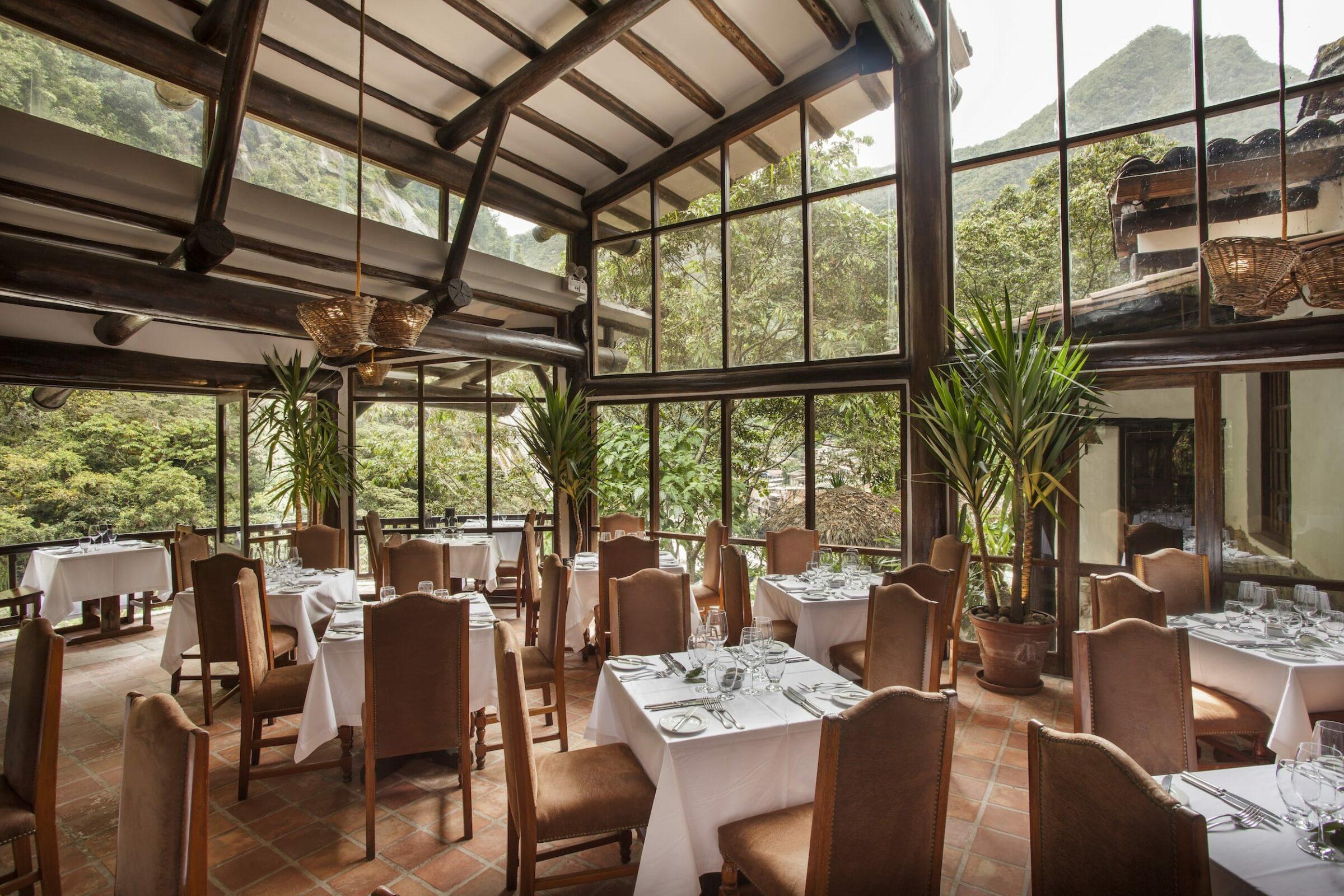 Elegantes Restaurantinterieur im Inkaterra Machu Picchu Pueblo Hotel mit großen Fenstern, die einen Blick auf die üppig grünen Berge bieten. Sonnendurchflutet, mit weißen Tischdecken, braunen Stühlen und hohen Pflanzen, die eine ruhige, einladende Atmosphäre schaffen.