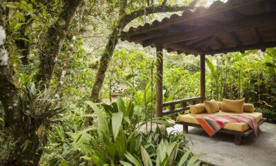 Ein gemütlicher Pavillon im Freien, eingebettet in üppiges Grün, verfügt über ein Holzsofa mit gelben Kissen und einer bunten gestreiften Decke, die eine ruhige, tropische Atmosphäre ausstrahlt. Inkaterra Suite im Inkaterra Machu Picchu Pueblo Hotel