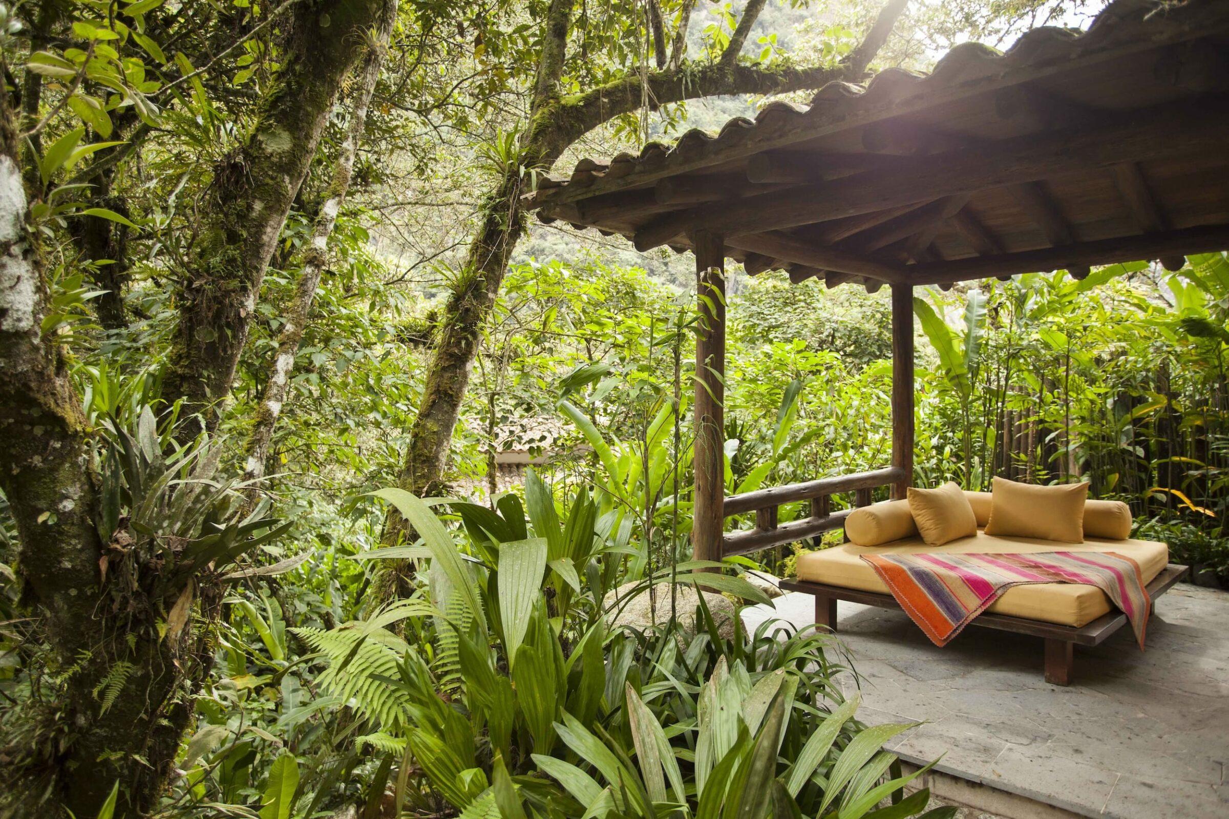 Ein gemütlicher Pavillon im Freien, eingebettet in üppiges Grün, verfügt über ein Holzsofa mit gelben Kissen und einer bunten gestreiften Decke, die eine ruhige, tropische Atmosphäre ausstrahlt. Inkaterra Suite im Inkaterra Machu Picchu Pueblo Hotel
