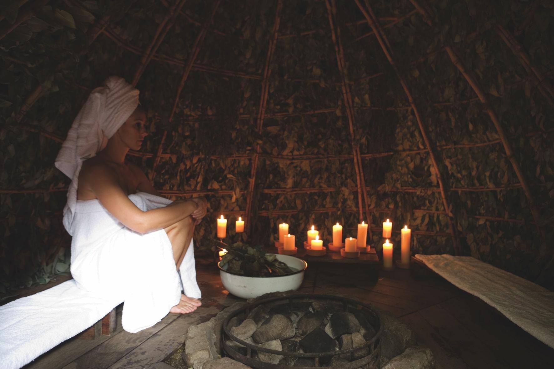 Eine Person, in ein weißes Handtuch gehüllt, sitzt meditativ in einer schwach beleuchteten, rustikalen Andensauna. Kerzen erhellen den Raum und schaffen eine ruhige, beruhigende Atmosphäre.