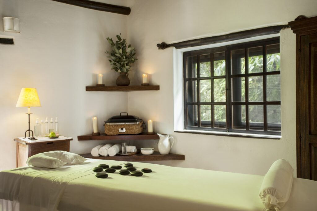 Gemütlicher Wellnessraum im Inkaterra Machu Picchu Pueblo Hotel mit einer Massageliege, die mit heißen Steinen geschmückt ist. Sanftes Licht von Kerzen und einer Lampe schafft eine ruhige Atmosphäre. Auf einem Regal liegen Handtücher, Öle und Dekorationsartikel.