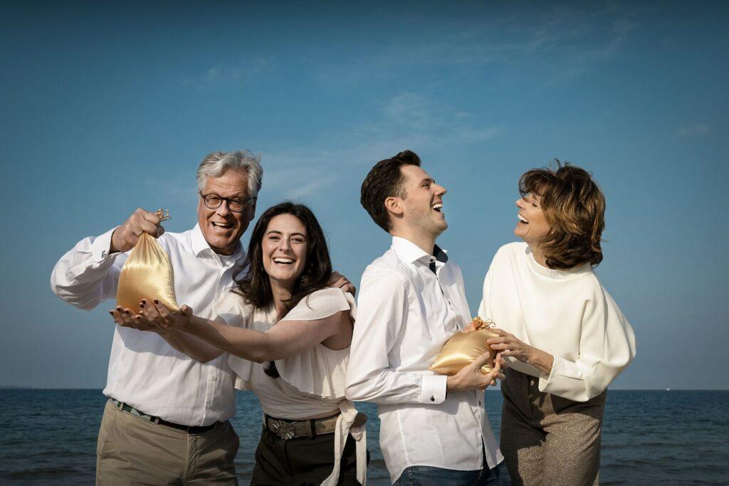 Die vierköpfige Inhaberfamilie des SANDnature steht fröhlich an einem Strand und hält jeweils einen goldenen Beutel in der Hand. Der Himmel ist klar, was die fröhliche und festliche Stimmung noch verstärkt.