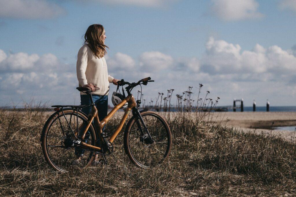 Eine Frau in einem weißen Pullover steht neben einem myBoo Bambusfahrrad auf einer grasbewachsenen Düne und blickt unter einem teilweise bewölkten Himmel auf das ruhige Meer.
