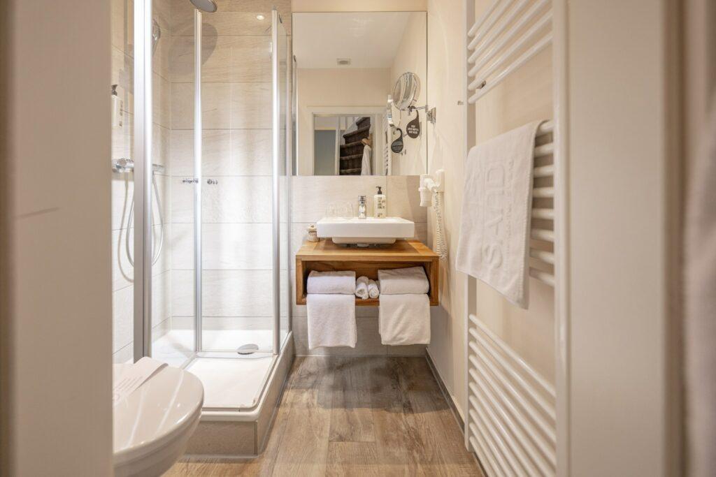 Modernes Badezimmer im SANDnature mit Glasdusche, wandmontiertem Waschbecken und Holzregal. Weiße Handtücher und Toilettenartikel sind ordentlich angeordnet. Helles und minimalistisches Design.