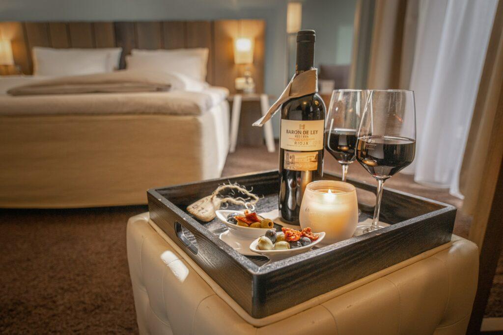 Gemütliches Hotelzimmer des Adults-Only-Hotels SANDnature an der Ostsee mit einem Tablett, auf dem eine Weinflasche, zwei Gläser Rotwein, eine brennende Kerze und kleine Schälchen mit Oliven und sonnengetrockneten Tomaten stehen. Warme Atmosphäre.