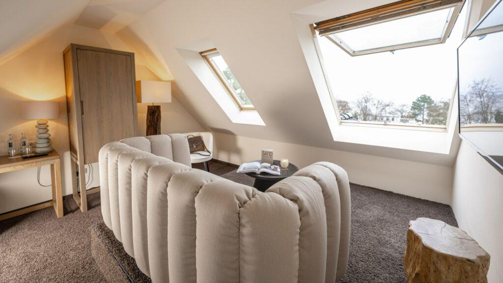 Gemütliches Wohnzimmer im Dachgeschoss des SANDnature Lifestylehotels mit beigem Sofa, braunem Teppich und Holzelementen. Durch die Oberlichter strömt natürliches Licht herein und schafft eine ruhige, einladende Atmosphäre.