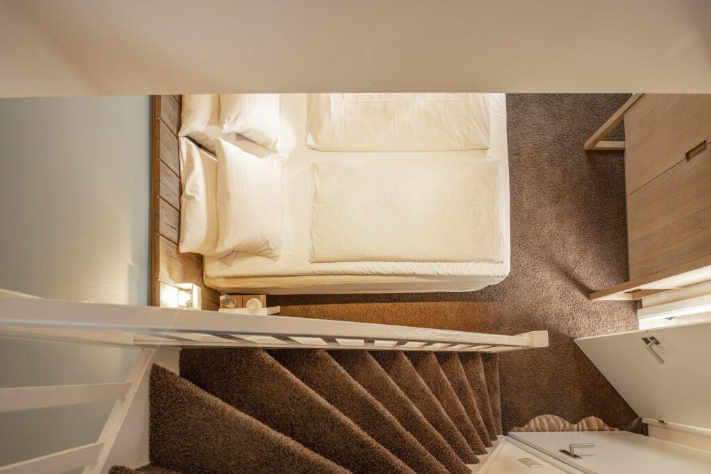 Blick auf ein gemütliches Schlafzimmer im Hotel SANDnature, das ausschließlich Erwachsenen vorbehalten ist, mit einem cremefarbenen Bett neben einer mit Teppich ausgelegten Treppe. Sanfte Beleuchtung und minimalistisches Dekor schaffen eine warme, einladende Atmosphäre.