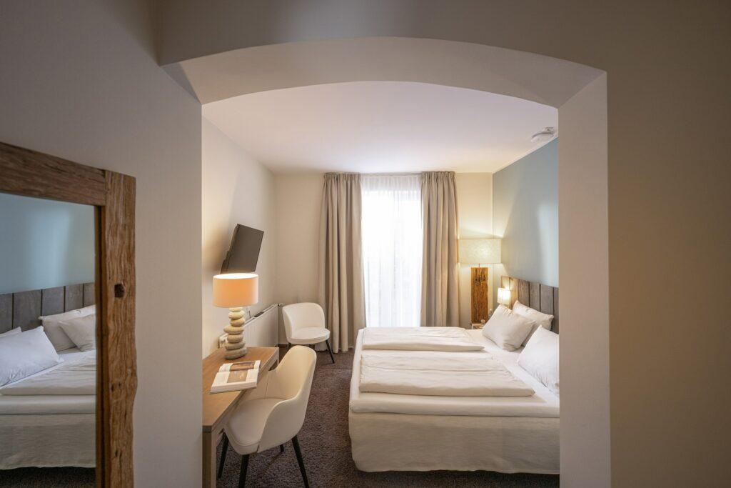 Modernes Hotelzimmer im SANDnature mit zwei ordentlich gemachten Betten, sanfter Beleuchtung durch eine Lampe, einem an der Wand montierten Fernseher und einem Schreibtisch mit Stuhl. Ein Fenster mit Vorhängen sorgt für natürliches Licht.