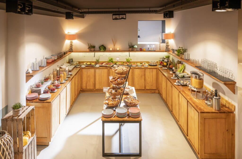 Helles Frühstücksbuffet mit Holzschränken, gefüllt mit einer Auswahl an Gebäck, Obst und Getränken. Auf dem Tisch in der Mitte wird Brot präsentiert. Warme, einladende Atmosphäre.