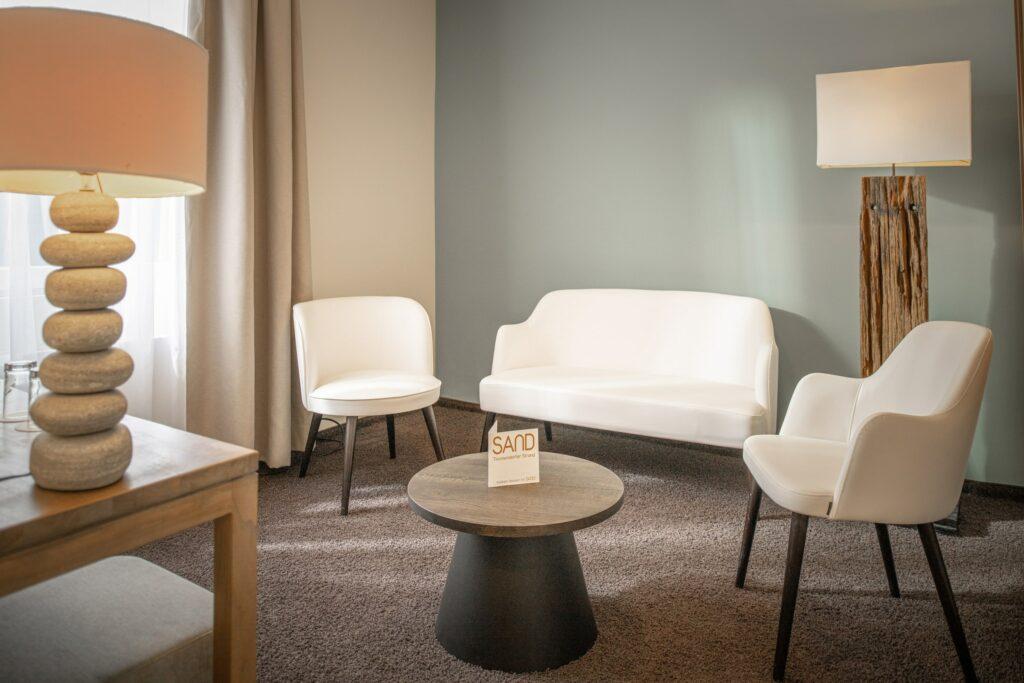 Ein gemütlicher Raum bei SANDnature mit weißen Stühlen, einem Sofa und einem runden Tisch auf einem Teppich. Sanftes Licht von einer Steinlampe und einer hohen Stehlampe schafft eine ruhige Atmosphäre.