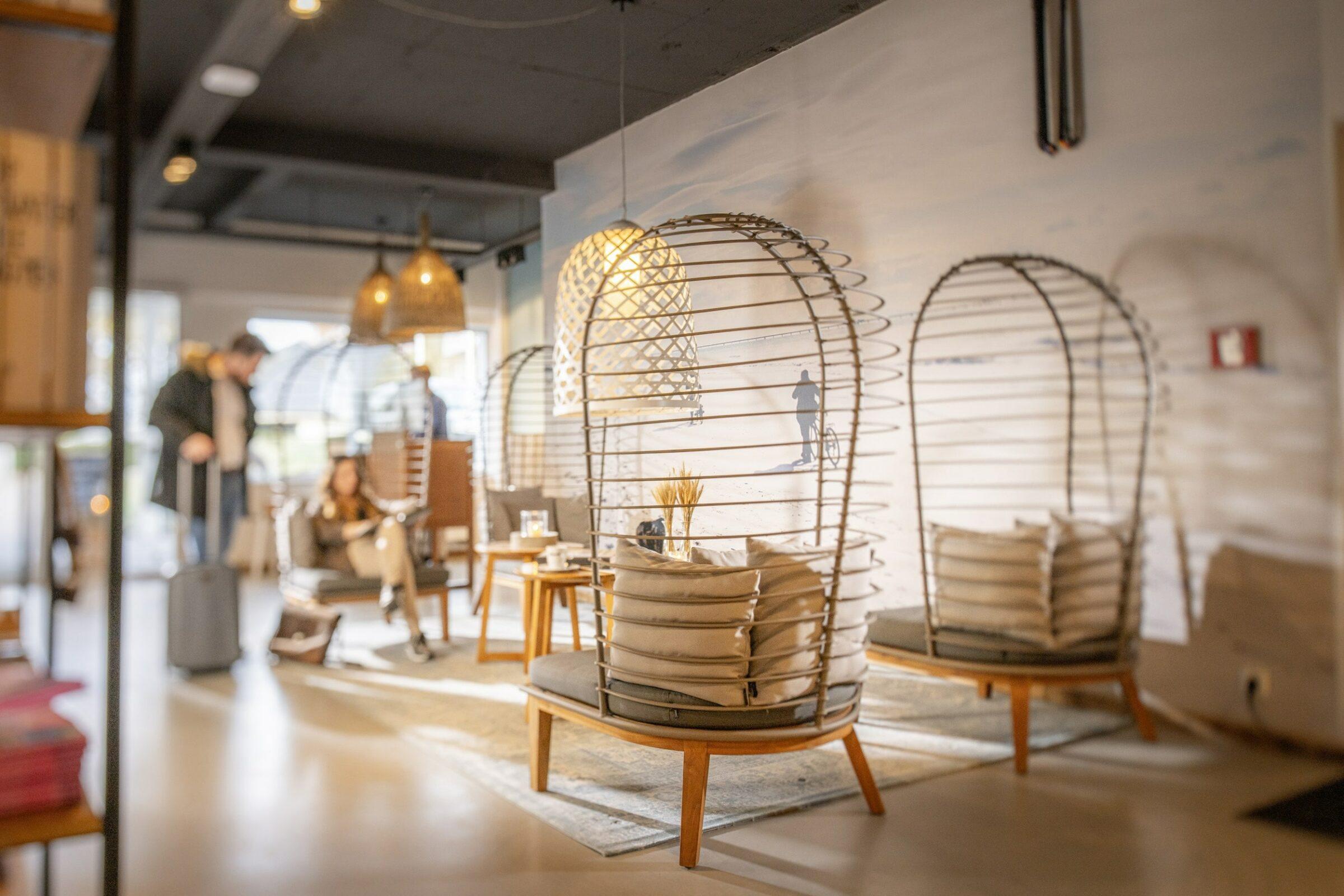 Eine gemütliche, moderne Lounge im nachhaltigen Hotel SANDnature nur für Erwachsene mit eiförmigen Korbstühlen, Kissen und kleinen Tischen. Sanfte Beleuchtung und verschwommene Figuren im Hintergrund schaffen eine entspannte Atmosphäre.