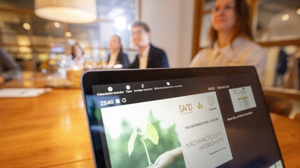 Nahaufnahme eines Laptop-Bildschirms, auf dem eine Präsentation zu einem Nachhaltigkeits-Workshop angezeigt wird. Im unscharfen Hintergrund sitzen drei Personen an einem Tisch.