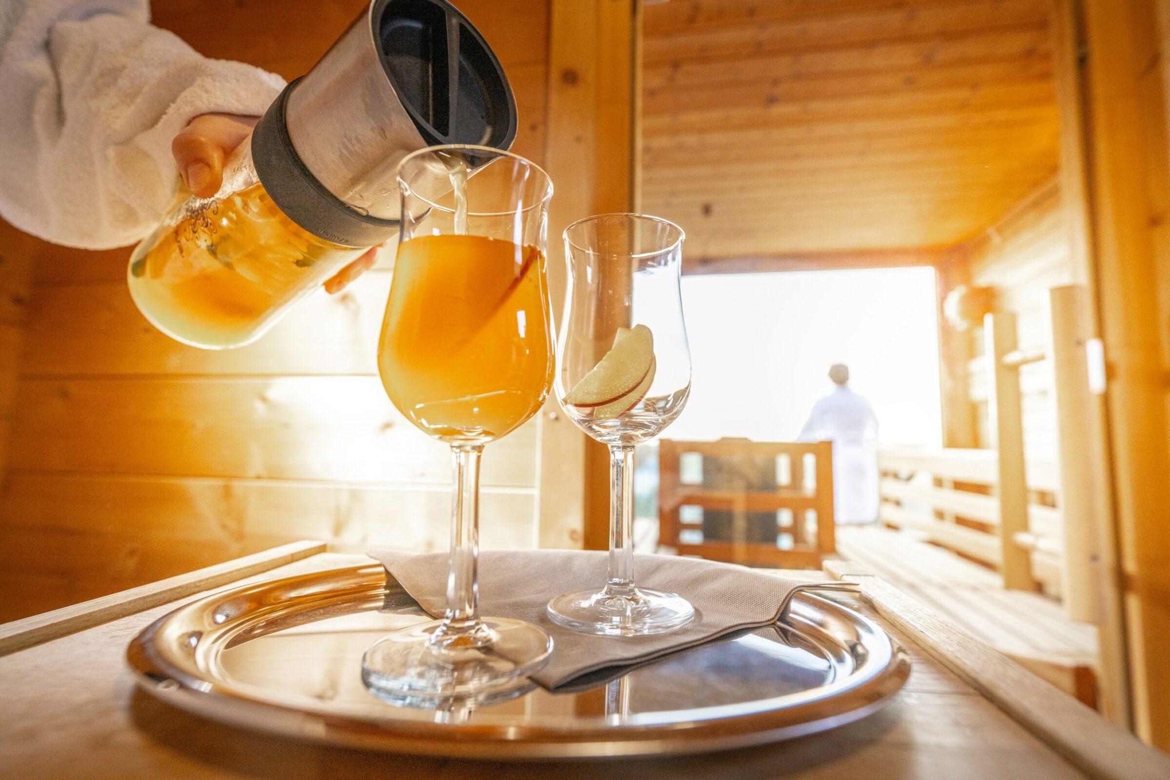 Eine Hand gießt Pfirsich-Eistee in ein Glas, neben einem weiteren Glas mit Pfirsichscheiben, auf einem Tablett in der gemütlichen Holzsauna auf dem Dach des Lifestylehotels SANDnature an der Ostsee. Im Hintergrund steht eine Person in einem weißen Bademantel.