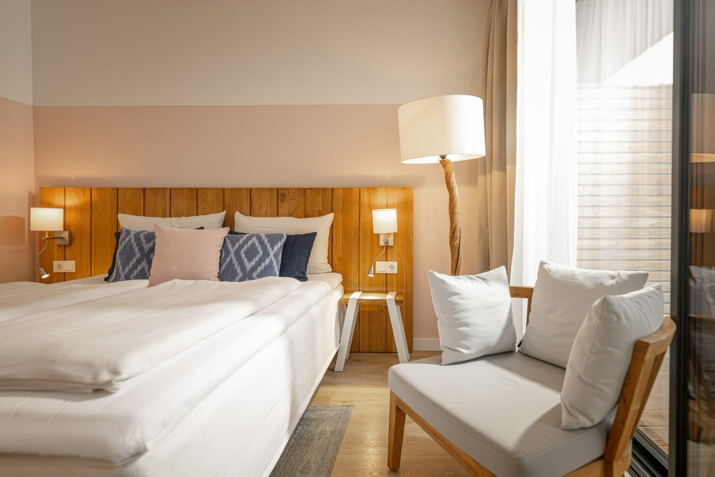 Ein ruhiges Schlafzimmer im Lifestylehotel SANDnature in Timmendorfer Strand, Deutschland, mit einem Kopfteil aus Holz, einem gemütlichen Doppelbett und weichen Kissen. Sanftes Licht von Lampen und ein großes Fenster schaffen eine ruhige, einladende Atmosphäre.