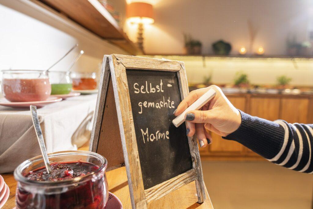 Eine Person schreibt auf eine kleine Tafel mit der Aufschrift „Selbstgemachte Marmelade” inmitten von Marmeladengläsern auf einem Tisch, wodurch eine gemütliche, hausgemachte Atmosphäre entsteht.