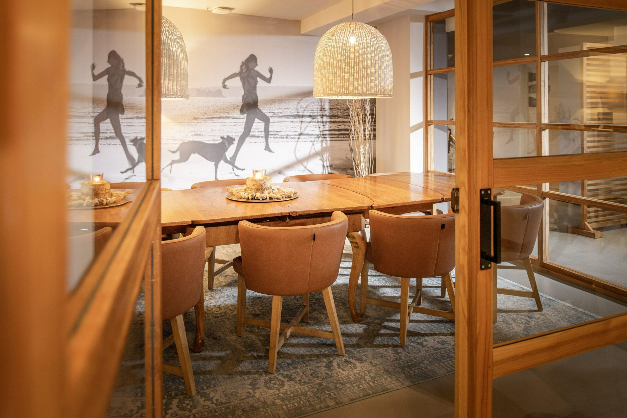 Gemütliches Esszimmer mit einem Holztisch, gepolsterten Stühlen und zwei Pendelleuchten. Die Wanddekoration zeigt eine Person, die mit einem Hund am Strand entlangläuft, und schafft eine warme, einladende Atmosphäre.