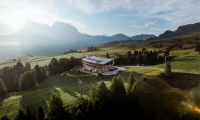 Luftaufnahme einer ruhigen Berglandschaft bei Sonnenaufgang, mit dem Paradiso Pure.Living Vegan Hotel inmitten grüner Felder und Bäume. Im nebligen Hintergrund erheben sich Berge.