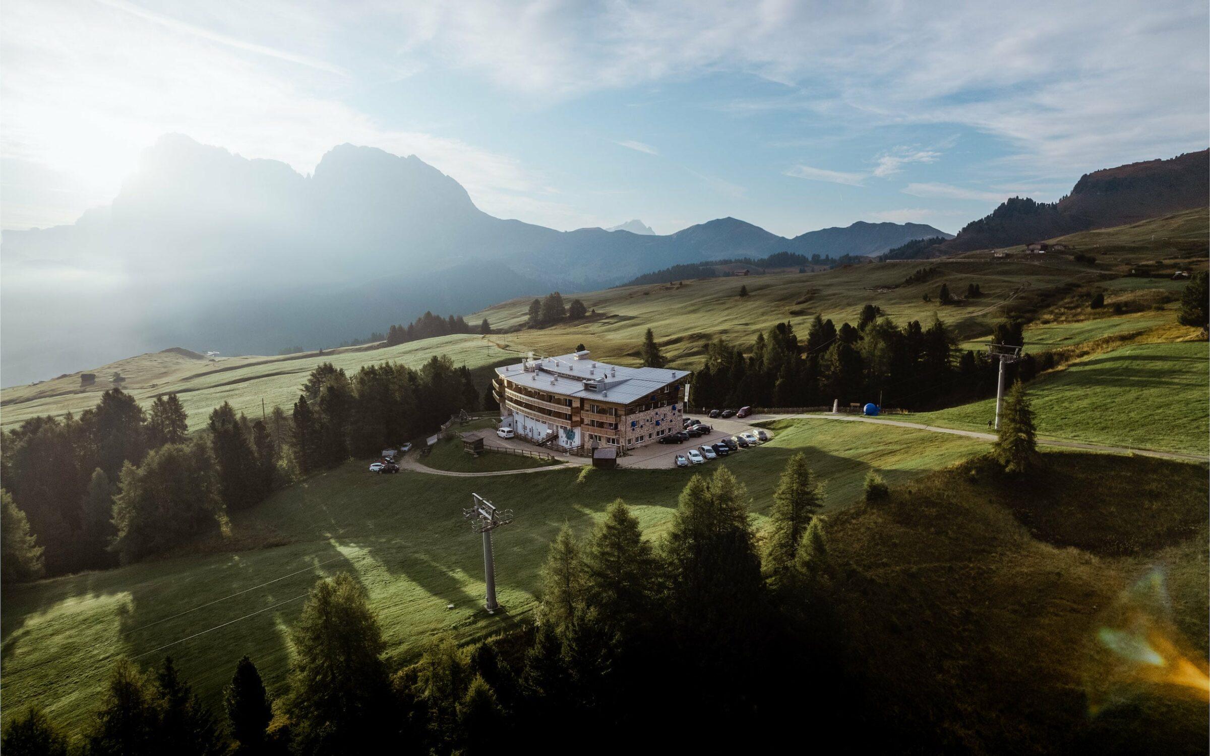 Luftaufnahme einer ruhigen Berglandschaft bei Sonnenaufgang, mit dem Paradiso Pure.Living Vegan Hotel inmitten grüner Felder und Bäume. Im nebligen Hintergrund erheben sich Berge.