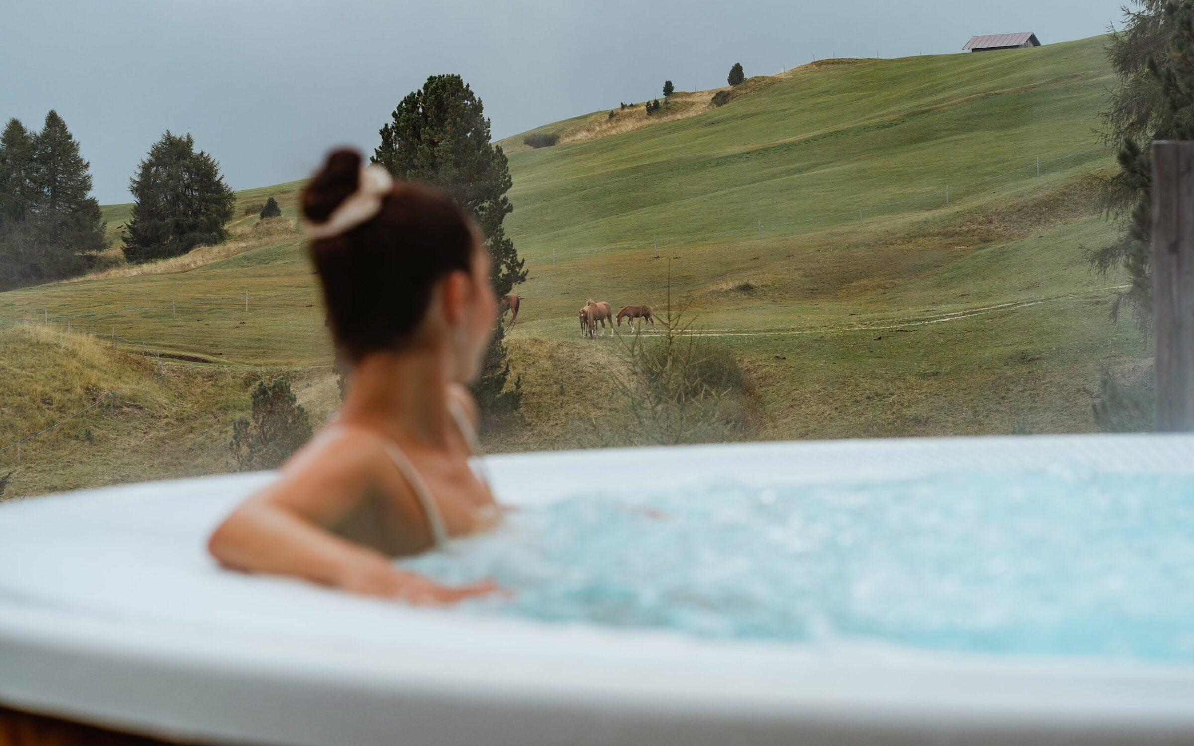 Eine Frau entspannt sich in einem Whirlpool und blickt auf eine ruhige, grüne Landschaft mit grasenden Pferden in der Ferne und einem bewölkten Himmel.